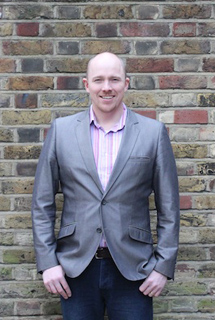 A smiling man wearing a grey blazer and a pink striped shirt stands before a brick wall.