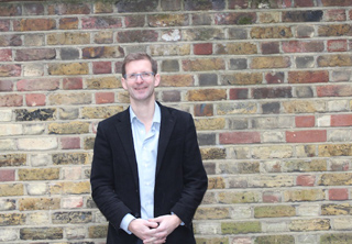 A man wearing glasses and a light blue shirt under a black blazer stands before a multi-colored brick wall.