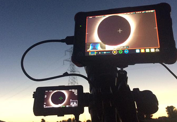A camera monitor on a tripod shows a dark solar eclipse against a bright sky at dusk.