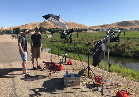 Two men stand by tripods, umbrellas, and lighting gear on a paved road near a field and stream.
