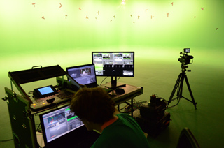 A person sits at a desk with multiple monitors and a video camera on a tripod in a green screen studio.