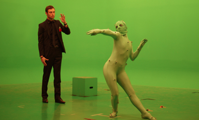 A man in a black suit watches a person in a green motion-capture suit posing against a green screen.