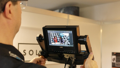 A person's hands holding a professional camera monitor displaying a scene of two people in a checkered room.
