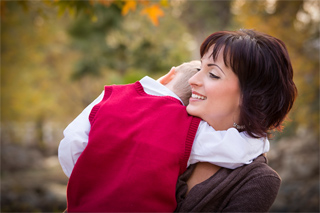 A smiling woman with dark hair embraces a child wearing a red sweater vest outdoors.