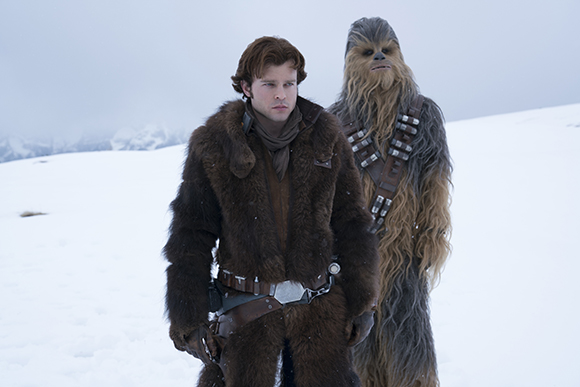 A man in a brown fur coat stands next to a tall, hairy creature in a snowy field.