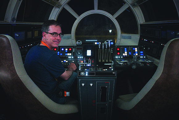 A man in a blue shirt sits in a pilot's seat facing a dark cockpit filled with glowing buttons and levers.
