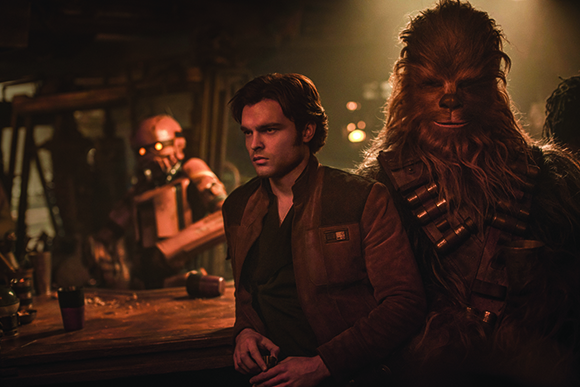 A man in a brown vest stands next to a tall, furry Wookiee in a dark, smoky room.