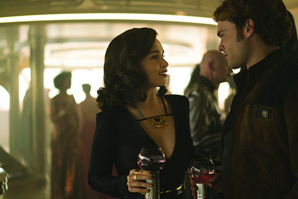 A woman in a black dress and a man in a dark jacket look at each other while holding wine glasses.