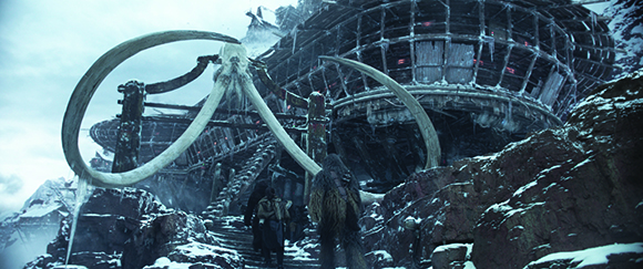 A large, circular metal structure built into a snowy mountain, framed by two massive mammoth tusks.