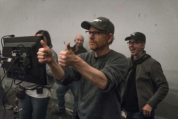 A man in a baseball cap and glasses gives two thumbs up while standing on a film set.