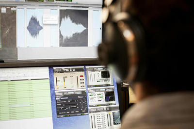 A person wearing headphones views audio waveforms and editing software on two computer monitors.