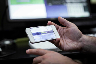 A person holds a white smartphone displaying the word 'VICTORY' in front of a blurred computer monitor.