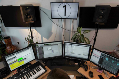 A desk with four computer monitors displaying audio software, a MIDI keyboard, and two studio monitors.