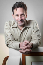 A man with dark hair and a light beard wearing a tan button-down shirt, leaning forward with hands clasped.
