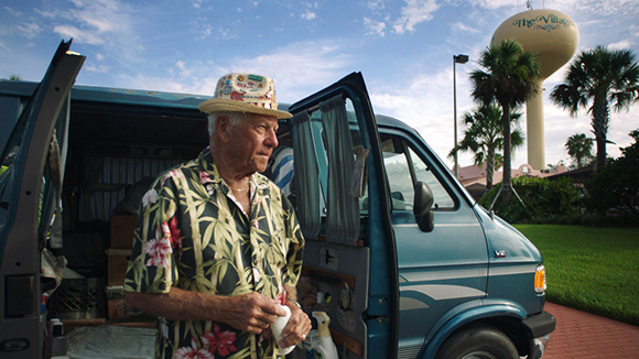 An older man wearing a straw hat and floral shirt stands next to an open blue van under a blue sky.