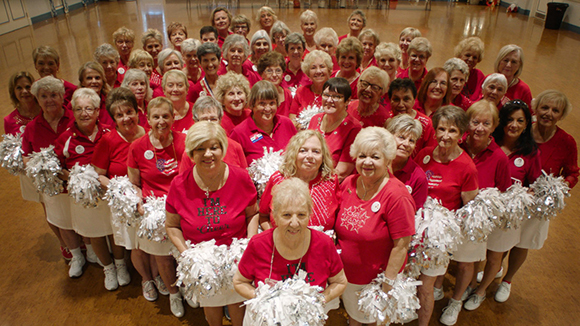 A large group of women wearing red shirts and holding silver pom-poms, posing together in a gymnasium.