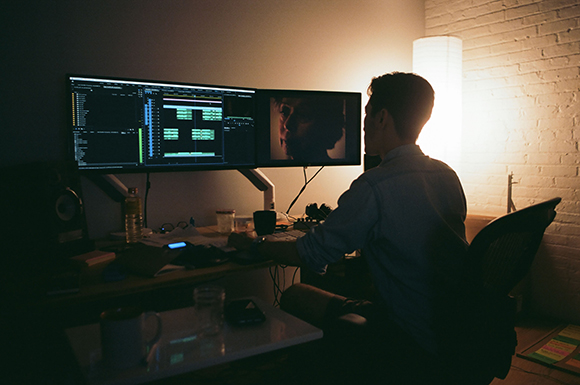 A person sits at a desk in a dark room, working on two computer monitors illuminated by a lamp.