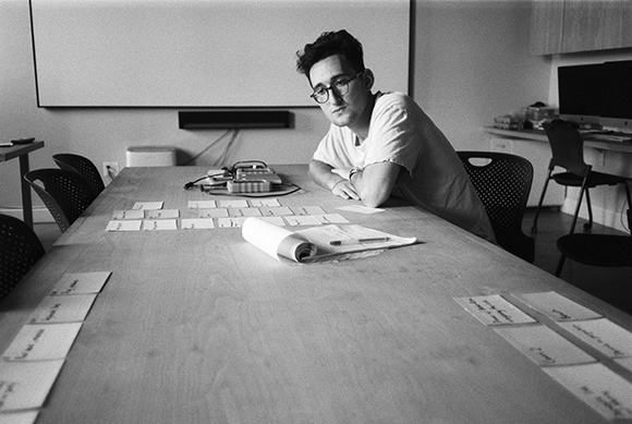 A man in glasses leans on a long wooden table covered with small white cards in a black and white photo.