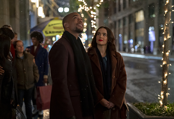 A man in a maroon coat and a woman in a brown coat look upward amidst city lights and a rainy street.