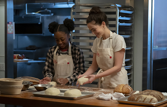 Two women in white aprons kneading dough on a flour-dusted wooden table in a commercial kitchen.