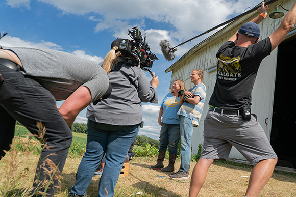A film crew uses a large camera and a boom microphone to film two women standing in a field.