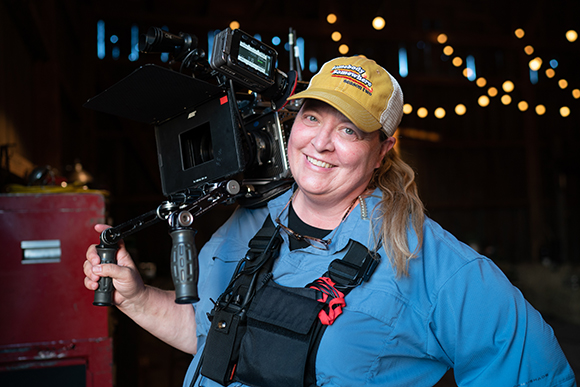 A smiling woman in a blue shirt and yellow cap carries a large professional video camera on her shoulder.