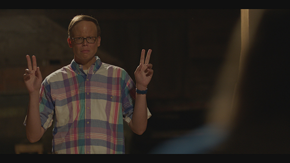 A man wearing glasses and a plaid shirt holds up two fingers in peace signs on both hands.