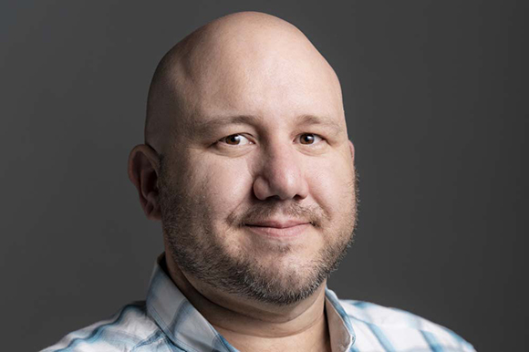 A bald man with a short beard and a light blue plaid shirt smiling slightly against a dark gray background.