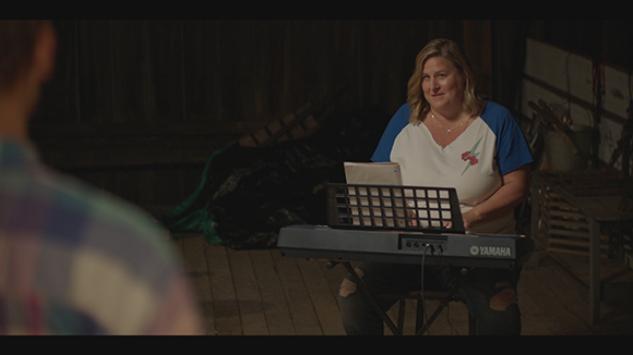 A woman in a white and blue shirt sits playing a Yamaha electronic keyboard in a dark room.