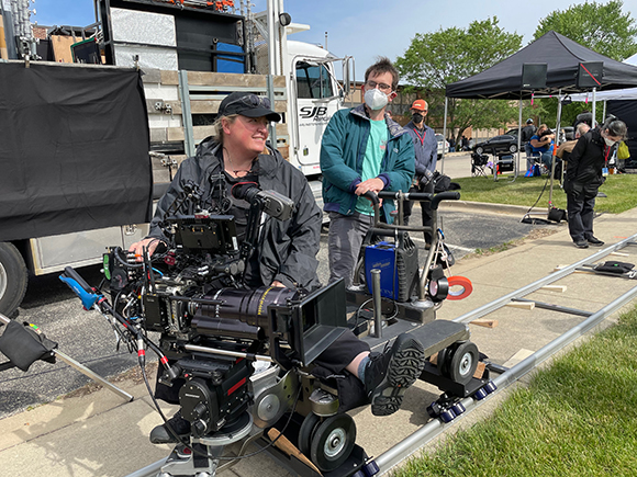 A camera operator and crew member stand near a professional cinema camera on tracks outdoors.