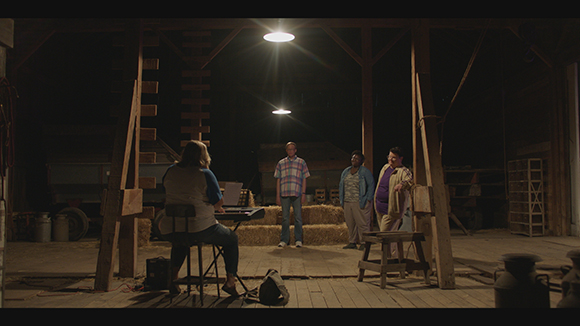 A person sits at a keyboard in a dark barn facing three people standing near hay bales under overhead lights.