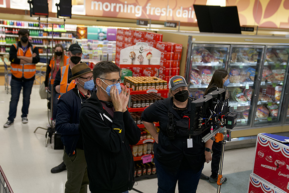 A film crew with a camera and crew members stands in a grocery store aisle near refrigerated cases.