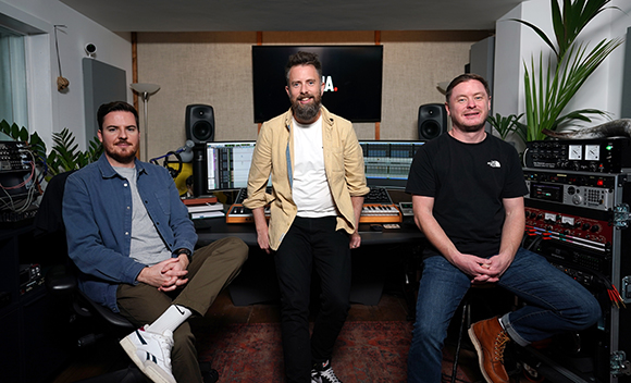 Three men pose in a music studio with mixing consoles, monitors, and audio equipment.