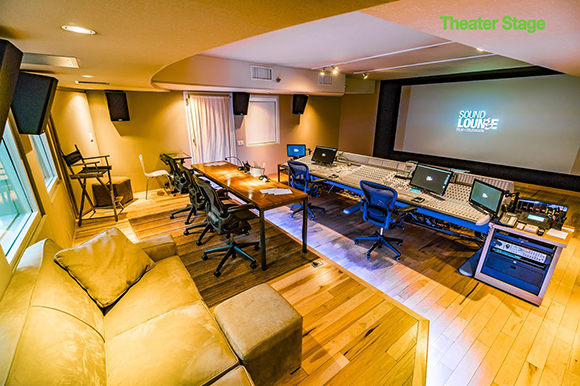 A professional audio mixing studio featuring a large console, multiple monitors, and a projection screen.