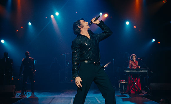 A man in a black sequined shirt sings into a microphone on a stage with blue and orange spotlights.