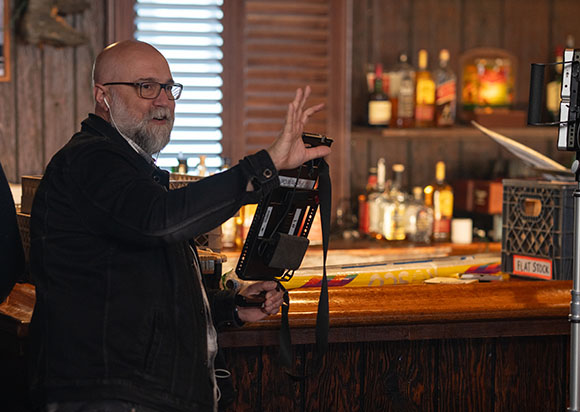 A bald man with glasses and a beard gestures with one hand while holding a camera accessory in a wooden bar.