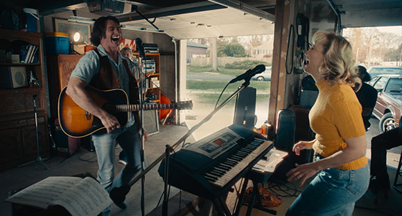 A man plays acoustic guitar while singing and a woman sings next to a keyboard in a garage.
