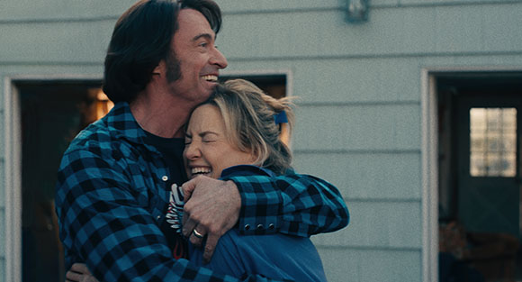 A man in a blue plaid shirt smiles while giving a blonde woman a tight hug.