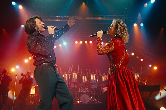A man and woman singing into microphones on a stage with bright, colorful spotlights.