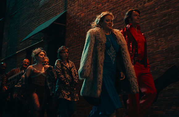 Four people in glamorous attire walk past a brick wall under dim lighting.