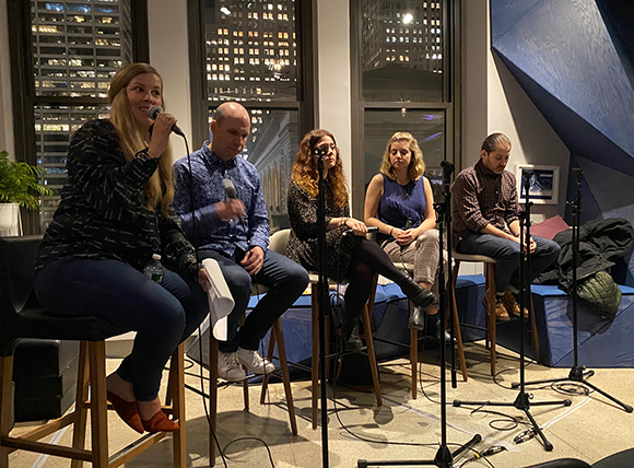 Five people sit on stools in a panel discussion, with microphones in front of them. City lights are visible through a window