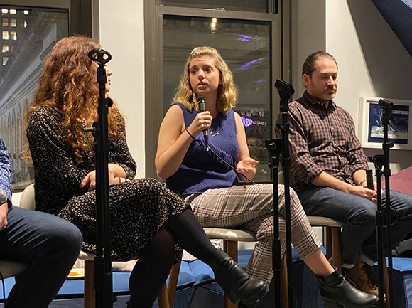 Three people are seated on chairs, facing forward. The person in the center is speaking into a microphone and gesturing with