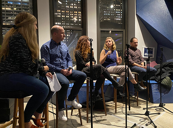 Five people sit on stools in a panel discussion. Microphones are in front of them. A city skyline is visible through the wind
