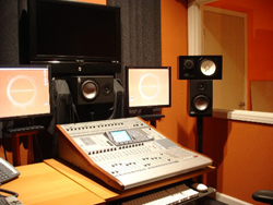 A mixing console sits on a desk between two computer monitors and studio speakers in a recording studio.