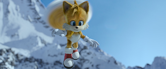 A yellow anthropomorphic fox with two tails flies through a bright blue sky over snowy mountains.