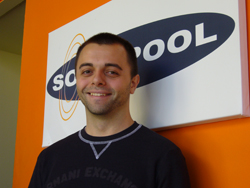 A smiling man in a black shirt stands in front of a white sign with a large blue oval logo.