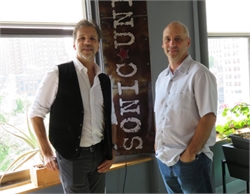 Two men standing side by side indoors next to a dark banner with white text.