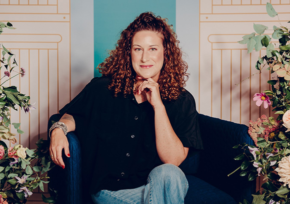A woman with curly hair sits in a blue armchair framed by floral arrangements against a pastel background.
