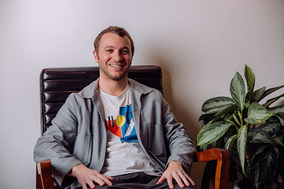 A smiling man in a grey jacket and graphic t-shirt sits in a black leather chair next to a leafy green plant.