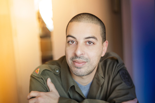 A man with short hair and light facial hair wearing an olive green military uniform.
